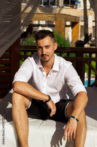 Urlaub in der Türkei. Hübscher gut aussehender jünger Mann, 20 Jahre alt sitzt auf dem Balkon, im Hintergrund ist weiße Gardine,. Er hat dunkle Haare, dunkler bart. Seine Hände sind auf den Knien