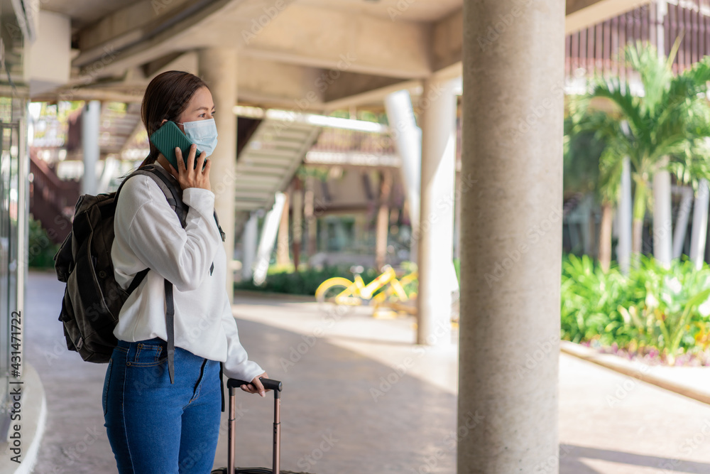 travel, woman in white long-sleeved shirt and jeans, wearing a mask.  using mobile phone during travel with suitcase in the left hand and backpack