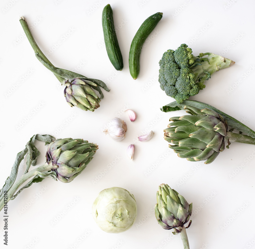 Many green vegetables - artichoke, broccoli, cucumber and kohlrabi placed around on white background with garlic. Concept very healthy and vitamin-rich food. Top view Many green vegetables - artichoke, broccoli, cucumber and kohlrabi placed around on white background with garlic. Concept very healthy and vitamin-rich food. Top view
