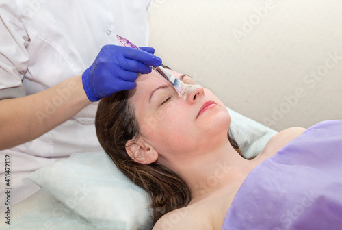Application of cosmetology mask on the face of a young woman at spa salon. Cosmetologist hand in blue protective glove touching patient face with brush..