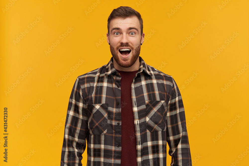 Fototapeta premium Surprised handsome young man in checkered shirt with beard and opened mouth looks amazed and shouting over yellow background