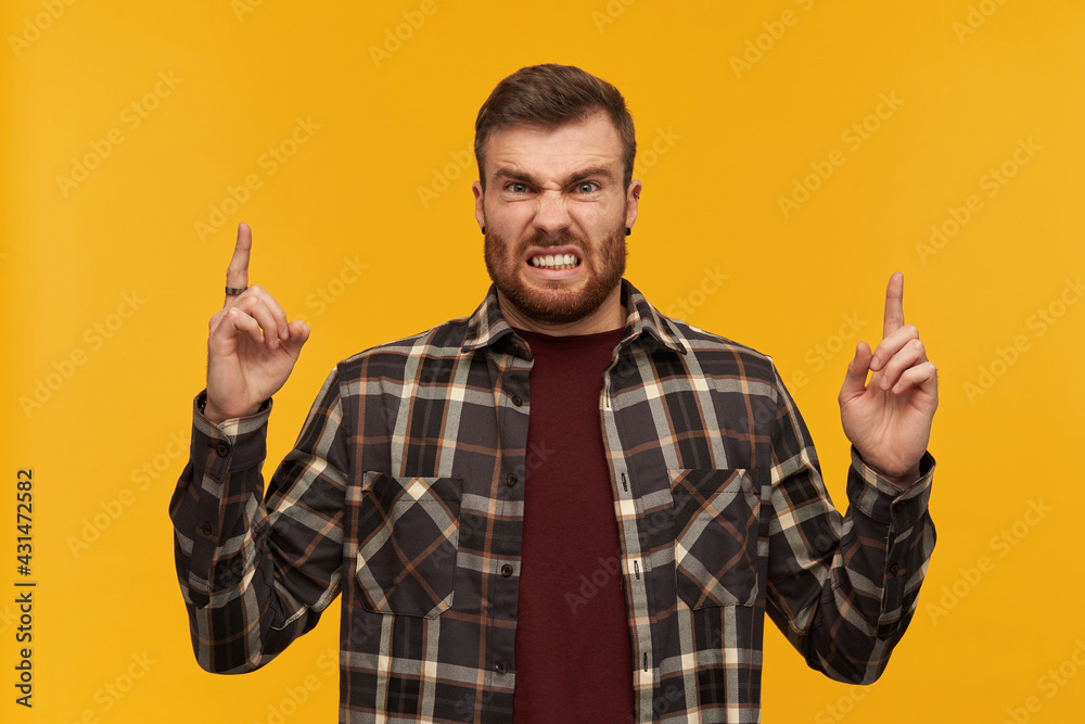 Mad furious young man in plaid shirt with beard pointing up to the sky by both hands and looking at camera over yellow background