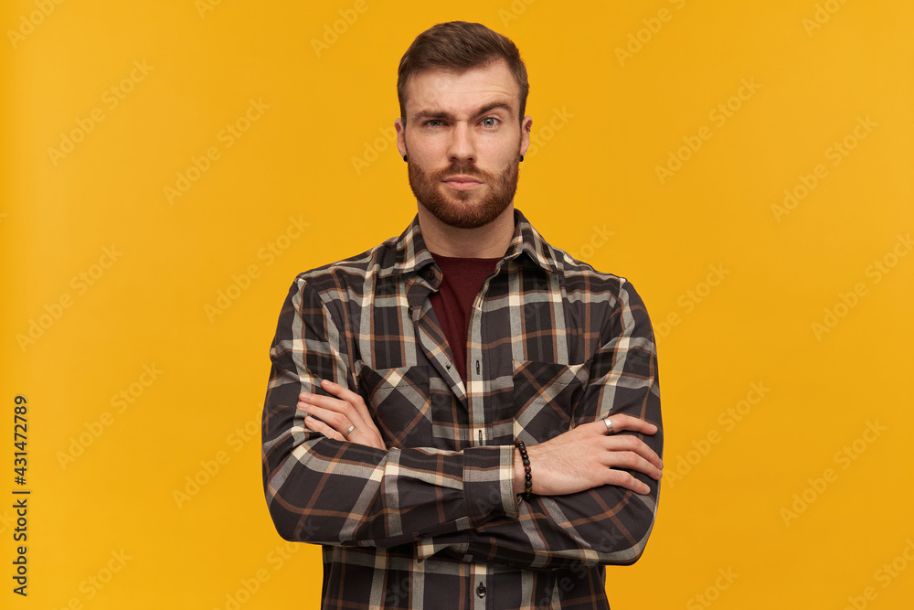 Serious attractive young bearded man in plaid shirt looks confident standing with hands folded and raised brow over yellow background Serious attractive young bearded man in plaid shirt looks confident standing with hands folded and raised brow over yellow background