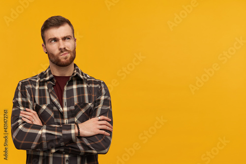 Thoughtful sceptical young man in checkered shirt with beard keeps arms crossed and thinking over yellow background Looking away to the side