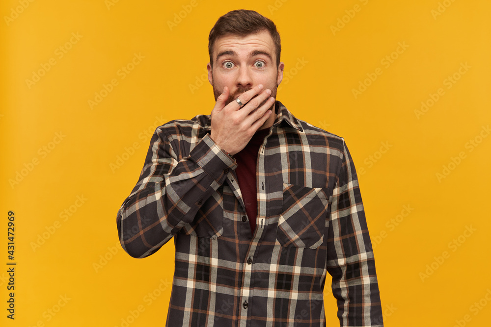 Obraz premium Scared shocked young man in plaid shirt with beard covered his mouth by hand and looks stunned over yellow background