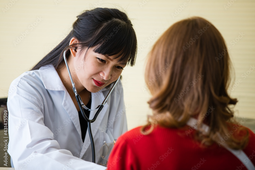 Doctor with long black hair white lab coat sitting checking heartbeat of lady who has blond hair wear red shirt in hospital healthcare center before discussing for recovering process. Doctor with long black hair white lab coat sitting checking heartbeat of lady who has blond hair wear red shirt in hospital healthcare center before discussing for recovering process.