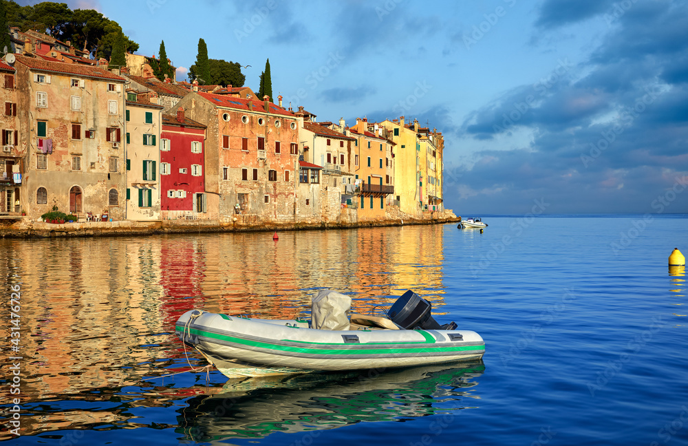 Rovinj, Istria, Croatia. Sunset in medieval old village town of Rovigno, colorful houses and boat in the harbor during morning sunrise. Picturesque scenic sky with clouds and blue water reflections.