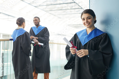 Group of young people wearing graduation gowns indoors in modern university interior, focus on smiling Middle-Eastern woman holding diploma and looking at camera in foreground, copy space