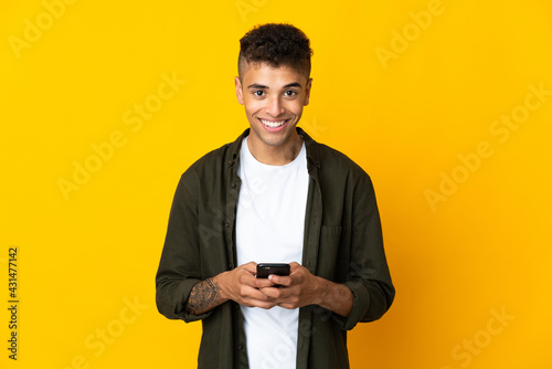 Young brazilian man isolated on yellow background looking at the camera and smiling while using the mobile
