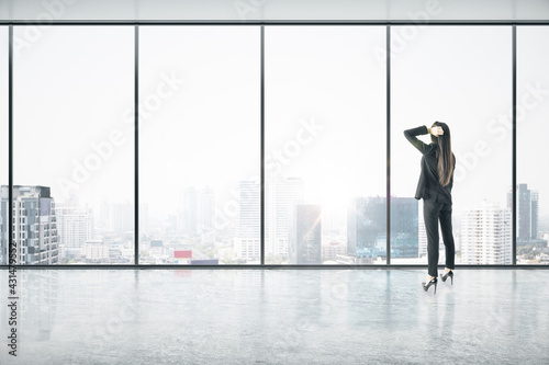 Businesswoman standing thinking in an empty big office room with big windows and city view. Real estate and future success concept
