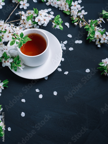 A white cup of tea on a stone table , branches of a blooming white cherry and petals . Copy space vertical photo postcard