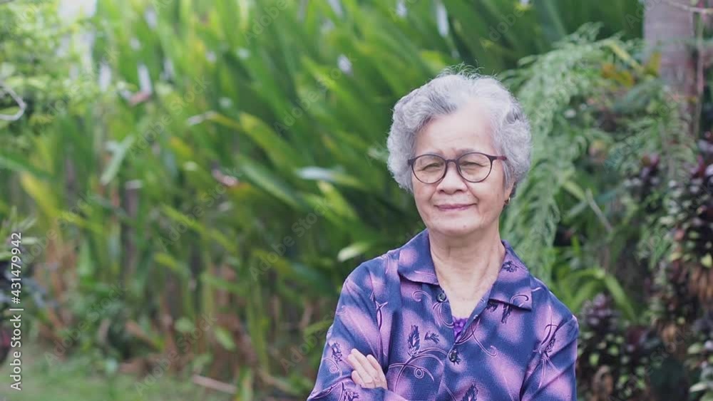 Portrait of a happy senior woman with short gray hair, wearing glasses, smiling, arms crossed, and looking at the camera while standing in a garden. Concept of aged people and relaxation. Portrait of a happy senior woman with short gray hair, wearing glasses, smiling, arms crossed, and looking at the camera while standing in a garden. Concept of aged people and relaxation.