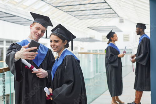 Two happy young people wearing graduation gowns and holding smartphone while taking selfie indoors in college interior