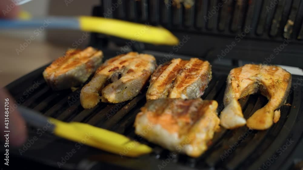 The man opens the electric grill with his hand and uses a spatula to remove the cooked trout fish from the baking sheet of the home grill. Close-up shot of ready-made grilled fish fillet meat The man opens the electric grill with his hand and uses a spatula to remove the cooked trout fish from the baking sheet of the home grill. Close-up shot of ready-made grilled fish fillet meat