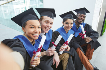 High angle view at diverse group of happy young people wearing graduation gowns and taking selfie outdoors