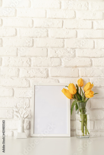 Fototapeta Naklejka Na Ścianę i Meble -  yellow tulips in a glass vase and blank photo frame on a white brick wall background