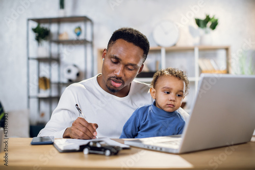 Focused young man working on laptop and writing on clipboard while sitting at table with baby boy on knees. African father taking care of his son and working from home.