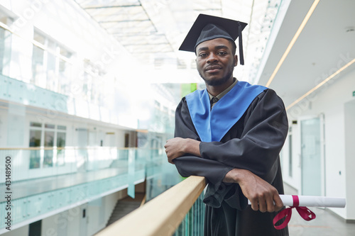 Waist up portrait of African-American young man wearing graduation gown and hat looking at camera while posing indoors