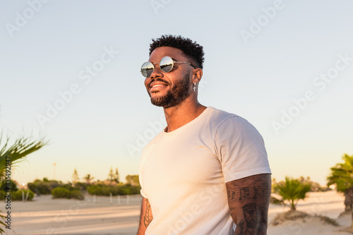 Portrait of a handsome black and young american man wearing sunglasses and smiling during sunset at the beach