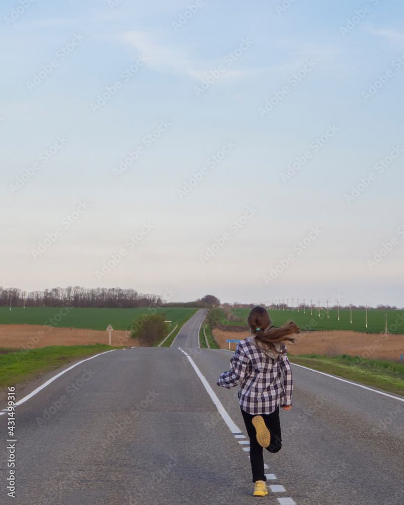 Obraz premium A young girl runs along the road. A girl with long blond hair runs away alone on a deserted road that goes into the distance among the fields. Healthy lifestyle.