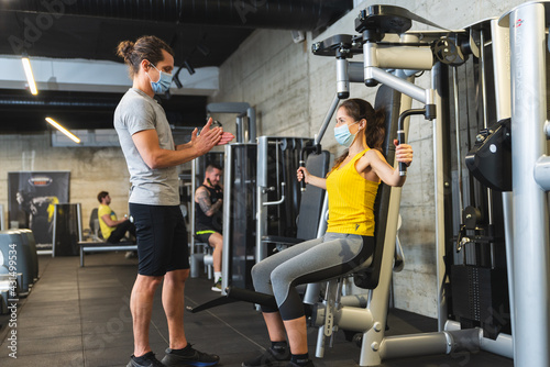 Personal trainer helping young female athlete in the gym while both of them wearing protective face masks