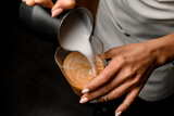 Close-up view on woman's hands pouring milk from jug into transparent glass of coffee drink