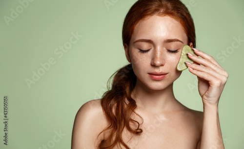 Skin care spa tcm. Redhead woman self massage facial skin with natural jade gouache guasha scraper. Girl uses gua sha scraper apply beauty cream or serum, rubbing skin, reduce puffiness dark circles