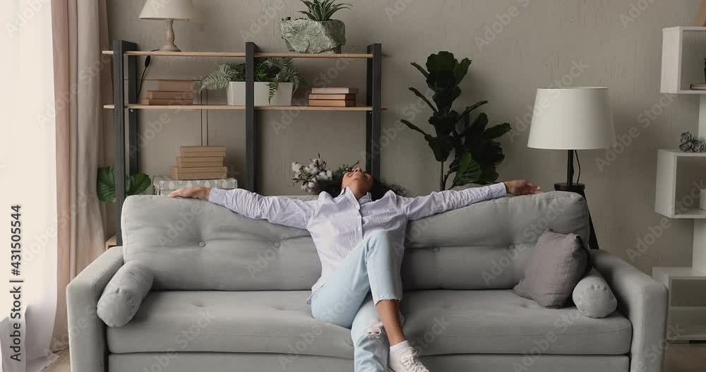 Cheery African woman sits on sofa take break in cozy living room put hands behind head enjoy carefree day off, breath fresh air inside of modern conditioned apartment. New day, happy homeowner concept