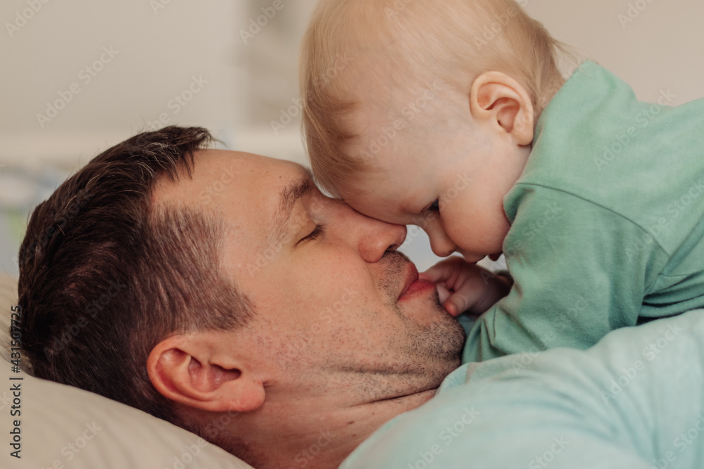 A young happy father lies with his baby son on the bed.Time together.Family concept.Father's Day concept.Paternal care.Newborn care.Close up,selective focus. A young happy father lies with his baby son on the bed.Time together.Family concept.Father's Day concept.Paternal care.Newborn care.Close up,selective focus.