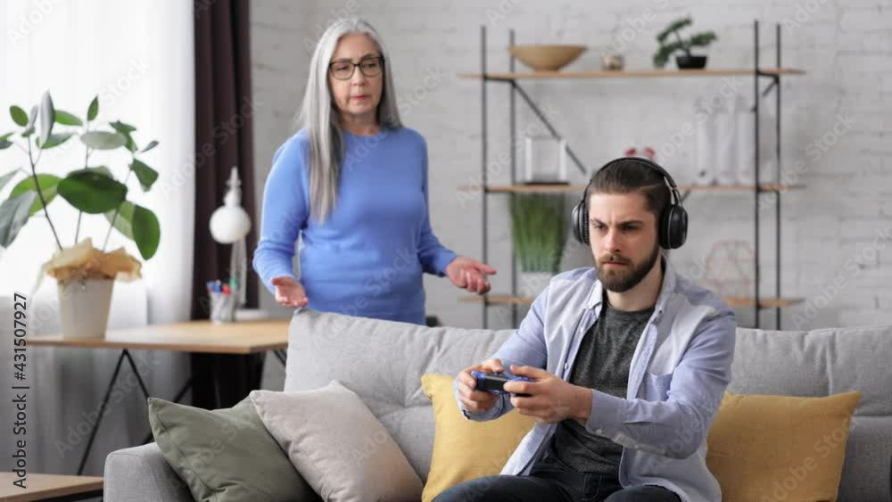Two generation family. Unemployed adult son playing video games at home using gamepads ignoring senior mother yelling at him. Video game addiction. Family and generations conflicts, misunderstanding. Two generation family. Unemployed adult son playing video games at home using gamepads ignoring senior mother yelling at him. Video game addiction. Family and generations conflicts, misunderstanding.