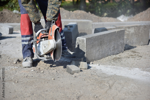 A worker picks up a circular saw from the sand, and there are concrete curbs nearby.