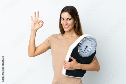 Young caucasian woman isolated on white background holding a weighing machine and doing OK sign