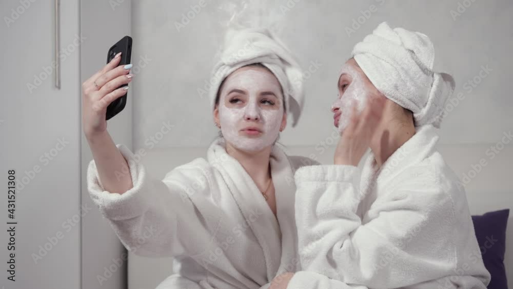 two girls in bathrobes and with clay masks on their faces take selfies sitting on the house on the bed. High quality 4k footage two girls in bathrobes and with clay masks on their faces take selfies sitting on the house on the bed. High quality 4k footage