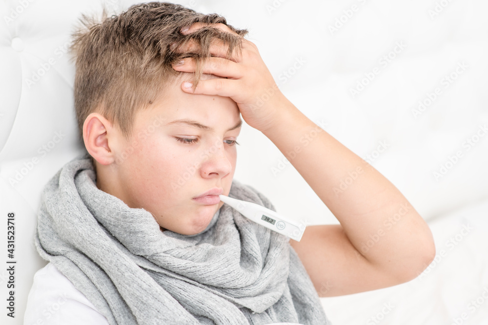 A sick little boy measures the temperature while lying on the bed at home. An unhealthy child holds his hand on his head in bead. Cold season.