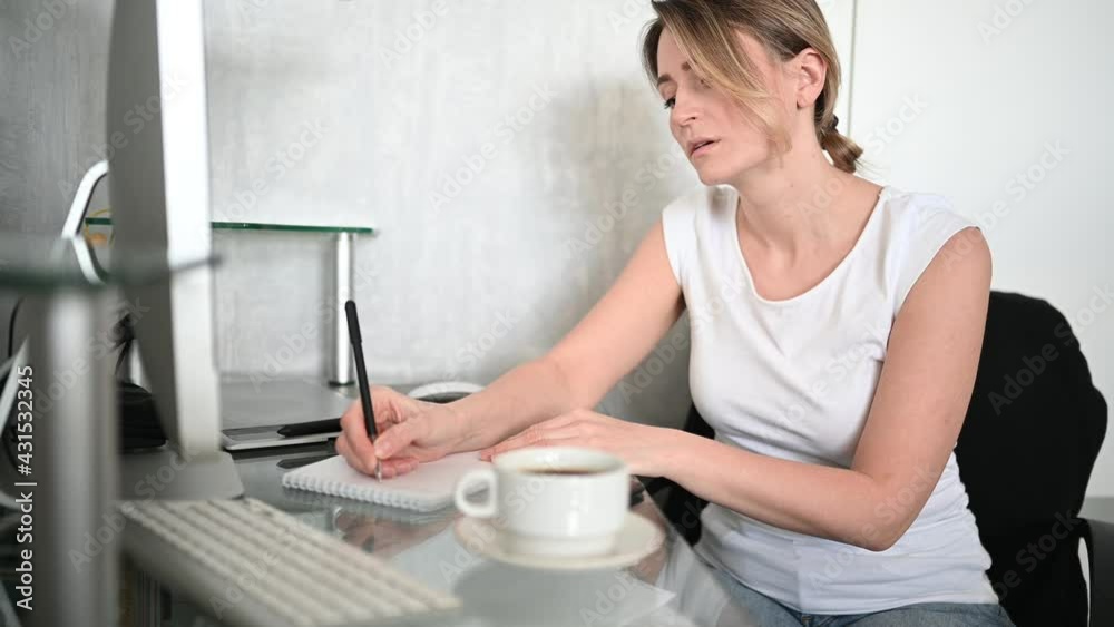 Young woman student sitting in front of computer with coffee learns online writes notes in notebook working at home office. Remote distance work education