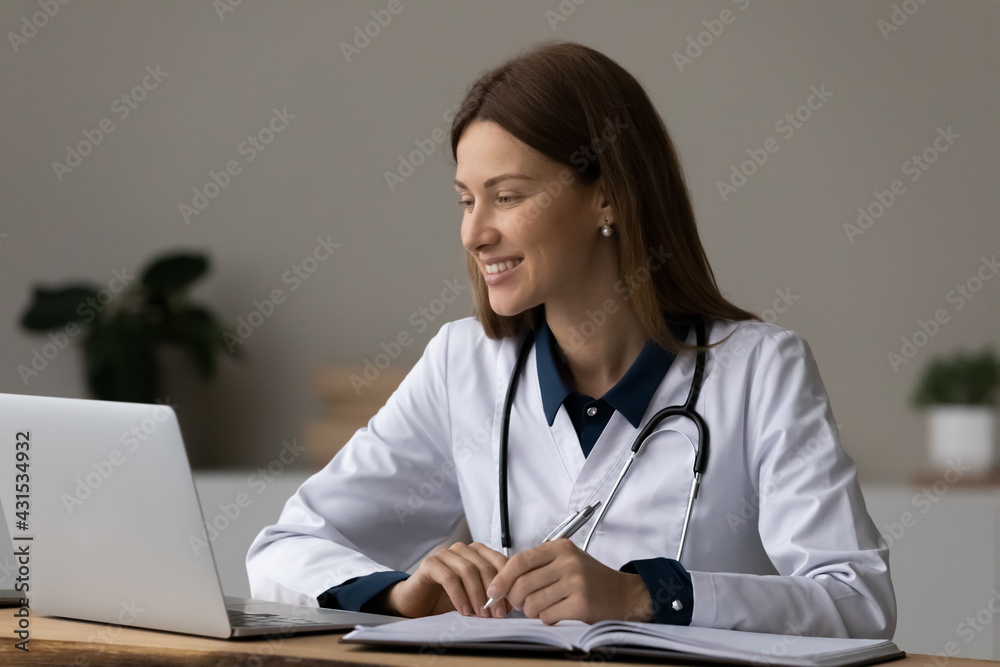 Smiling young female doctor in white medical uniform sit at desk in hospital work online on laptop. Happy woman GP or nurse consult patient client distant on computer in clinic. Healthcare concept. Smiling young female doctor in white medical uniform sit at desk in hospital work online on laptop. Happy woman GP or nurse consult patient client distant on computer in clinic. Healthcare concept.