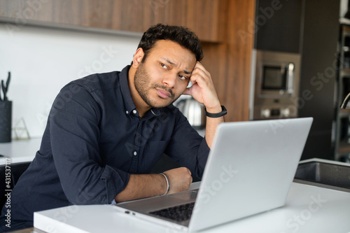 Depressed unemployed hindu man is using laptop for job seeking, upset pensive young indian guy sits in the kitchen at home with the laptop, rested head with hand, looks away, feels sad and worrying