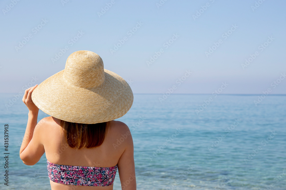 Back view of attractive fit young woman wearing bright bikini swimsuit with flower patterns and broad brim straw hat standing at the beach, enjoying ocean view. Background, close up, copy space