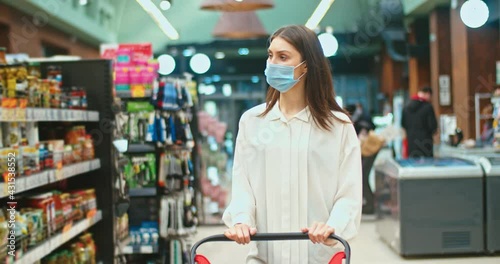 Successful housewife buying products, ingredients to cook dinner in supermarket. Adulr woman in mask choosing pickles and putting into shopping cart in grocery store. Virus, quarantine concept. photo