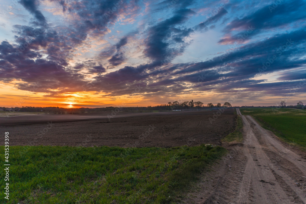 Sunset over rural meadow field and country road. Countryside landscape with path way under Scenic dramatic sky.