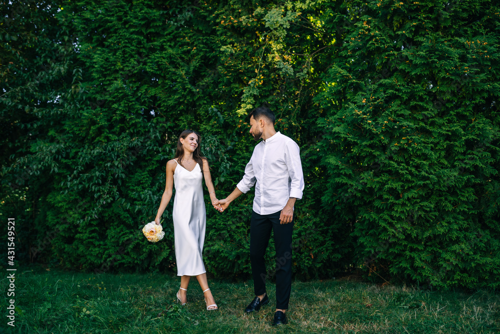 beautiful brunette bride in a white silky dress with her boyfriend walks in nature, holding a bouquet of flowers in her hands beautiful brunette bride in a white silky dress with her boyfriend walks in nature, holding a bouquet of flowers in her hands