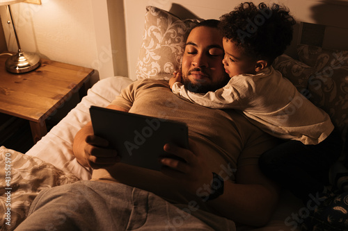 Little boy embracing his father while them lying on a bed. Young father reading book to his child from a digital tablet before sleeping. photo