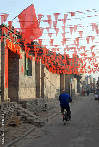 Pingyao in Shanxi Province, China: A backstreet in Pingyao with red flags flying above the road and an old man cycling. Pingyao old town is a famous ancient walled city in China.