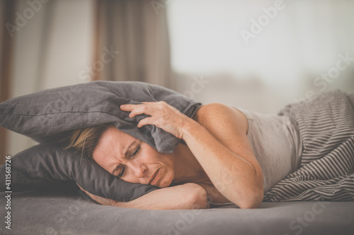 Sleepless lady covering ears with pillow. Noisy neighbors, tinnitus, insomnia or stress concept. Tired woman can't sleep. Awake in bed after coming home from her shift, work. photo