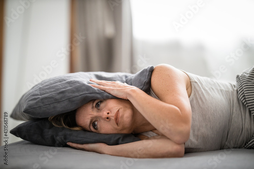 Sleepless lady covering ears with pillow. Noisy neighbors, tinnitus, insomnia or stress concept. Tired woman can\'t sleep. Awake in bed after coming home from her shift, work.