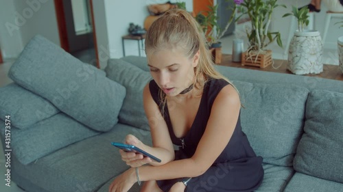 Gorgeous young blonde woman sitting on a sofa wearing sexy black dress with choker on her neck and using mobile phone looking at dating apps as she swipes and sends seductive messages