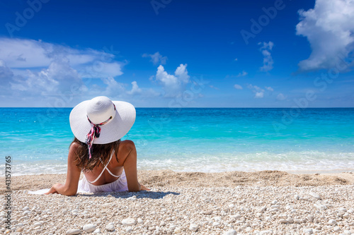 A tourist woman in a white dress and with hat on a beach enjoys the beautiful, turquoise Ionian Sea of Greece during her summer holiday © moofushi