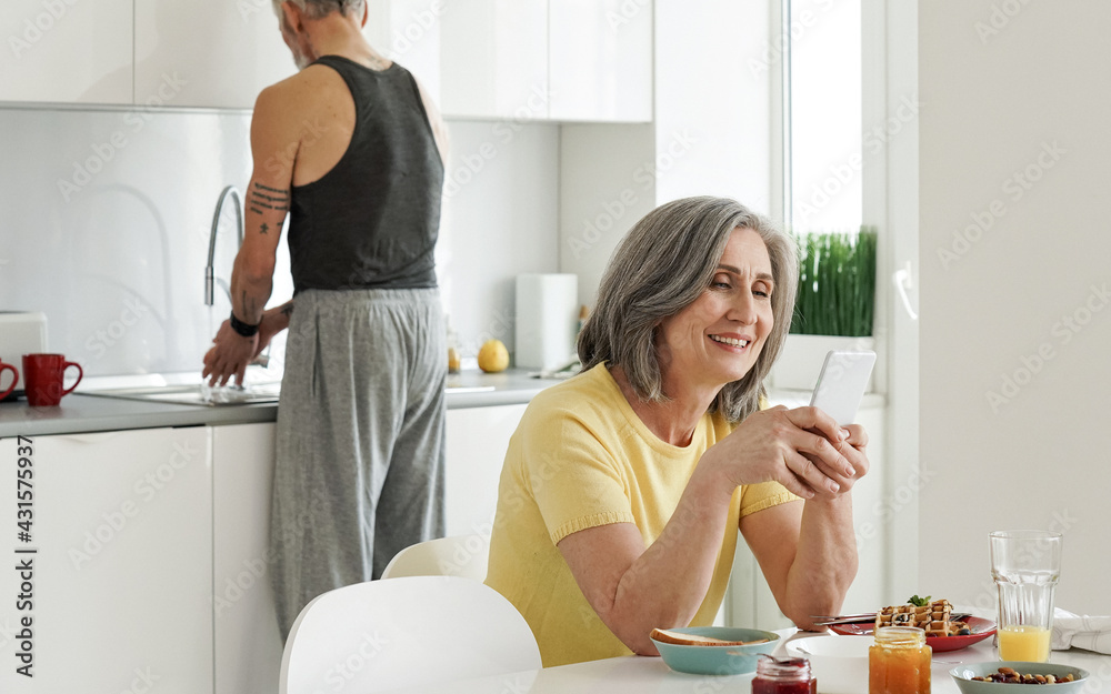 Happy older mature woman wife using mobile apps on cell phone ordering food delivery shopping in web store holding smartphone sitting at kitchen table while senior husband washing dishes at home. Happy older mature woman wife using mobile apps on cell phone ordering food delivery shopping in web store holding smartphone sitting at kitchen table while senior husband washing dishes at home.