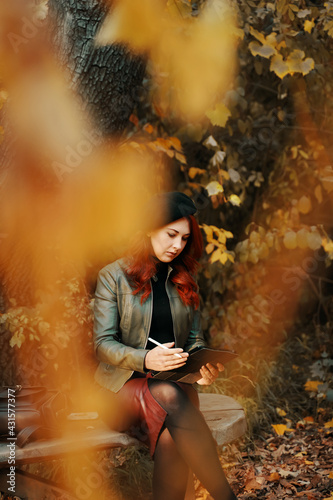 Pensive red-haired woman in the park on wooden bench. Pretty woman in beret and leather jacket. A person with a graphic tablet, a designer, a freelancer. Autumn portrait. © Konstantin Savusia