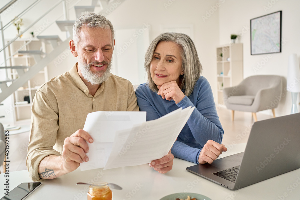 Middle aged senior old couple holding documents reading paper bills paying bank loan online, calculating pension fees, payments, taxes, planning family retirement money finances using laptop at home. Middle aged senior old couple holding documents reading paper bills paying bank loan online, calculating pension fees, payments, taxes, planning family retirement money finances using laptop at home.